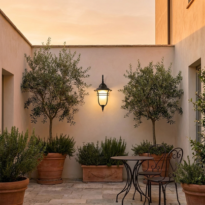 Traditional black lantern wall light glowing in plaster courtyard with olive planters at dusk